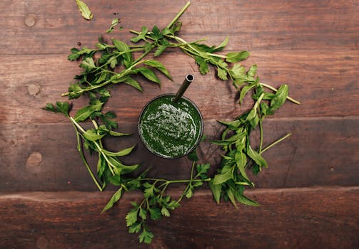 Top view of a green smoothie in a glass surrounded by fresh herbs on a wooden table.